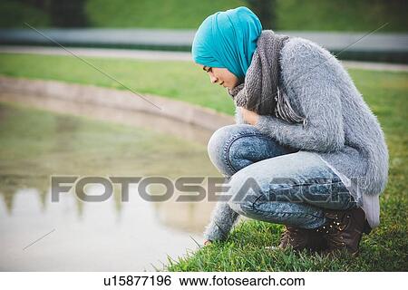Stock Photograph - Young woman wearing hijab crouching at park lakeside. Fotosearch