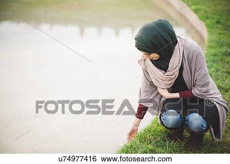 Stock Photograph - Young woman wearing hijab crouching next to park lake. Fotosearch