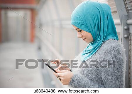 Stock Photo - Young woman wearing turquoise hijab using digital tablet on footbridge. Fotosearch