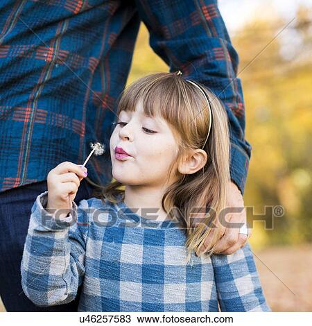 Stock Image - Girl (4-5) standing by father's side blowing off dandelion seed. Fotosearch
