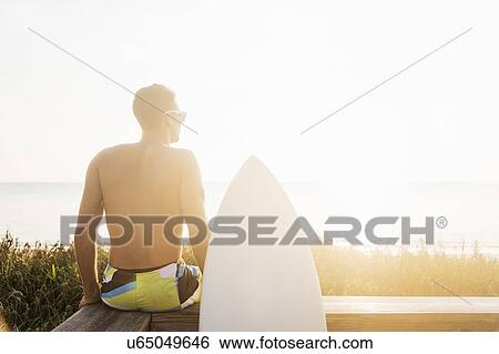 Stock Photograph - Rear view of surfer sitting on boardwalk. Fotosearch
