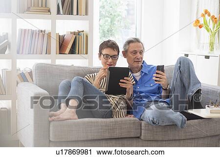 Stock Image - Senior couple sharing electronic devices on sofa. Fotosearch