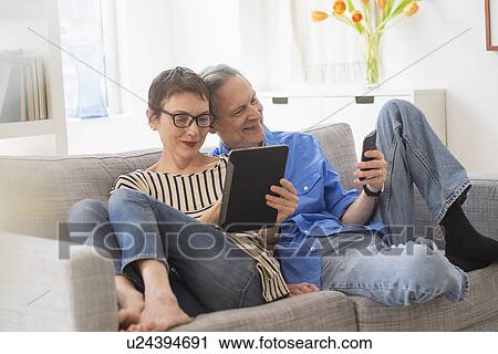 Stock Image - Senior couple sharing electronic devices on sofa. Fotosearch