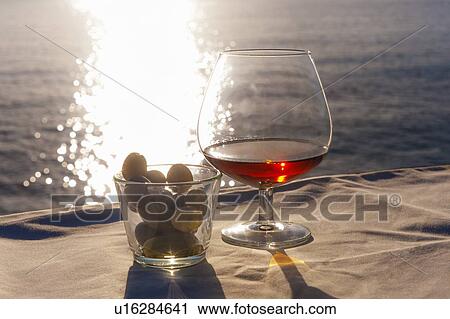 Stock Image - Brandy glass and olives on sunlit table at coast. Fotosearch