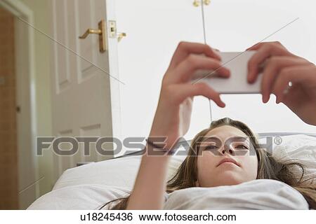 Stock Photograph - Girl lying on bed reading smartphone text messages. Fotosearch