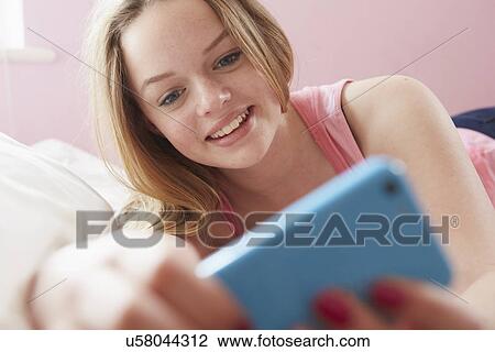 Stock Image - Girl lying on bed reading text message on smartphone. Fotosearch