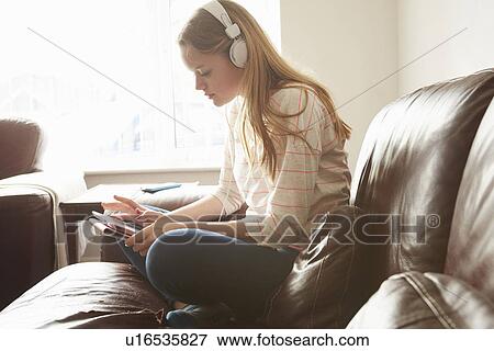 Stock Photo - Girl on sofa wearing headphones selecting music on digital tablet. Fotosearch