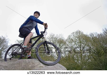 Stock Image - Low angle view of male mountain biker looking out from rock formation. Fotosearch