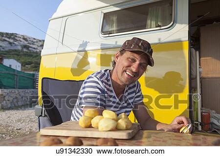 Stock Image - Man peeling potatoes beside van. Fotosearch