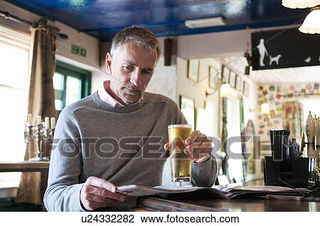 Stock Image - Mature man drinking beer and reading newspaper in pub. Fotosearch