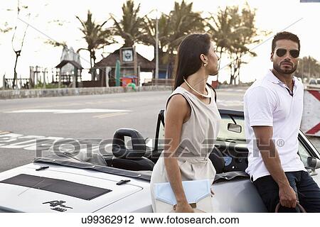 Stock Image - "Mid adult couple leaning on convertible car, by side of road". Fotosearch