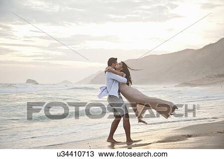 Stock Image - "Mid adult couple on beach, man lifting woman off ground, hugging". Fotosearch