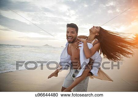 Stock Image - "Mid adult couple on beach, man carrying woman on back". Fotosearch