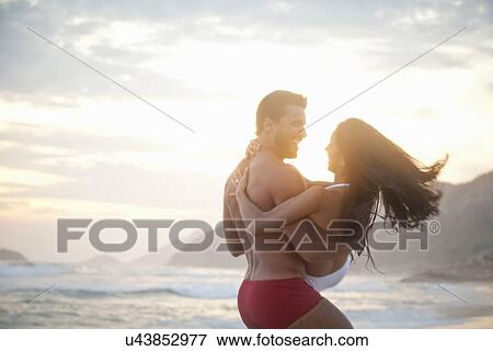 Stock Photo - "Mid adult couple on beach, man carrying woman in arms, rear view". Fotosearch