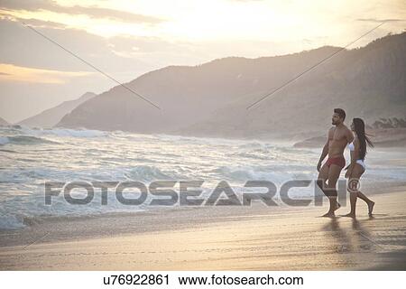 Stock Image - "Mid adult couple on beach, wearing swimwear, walking towards ocean". Fotosearch