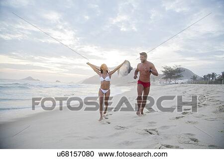 Stock Image - Mid adult couple running along beach in swimwear. Fotosearch