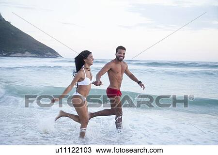 Stock Image - "Mid adult couple running along beach as tide comes in, hand in hand". Fotosearch
