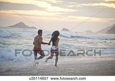 Stock Photo - "Mid adult couple running along beach as tide comes in, hand in hand, rear view". Fotosearch
