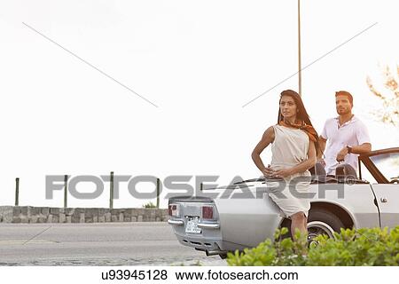 Stock Photo - Mid adult couple standing by convertible car by side of road. Fotosearch