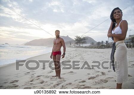 Stock Image - Mid adult couple standing on beach in swimwear. Fotosearch
