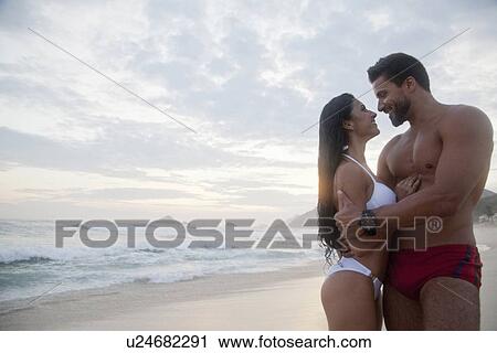 Stock Image - "Mid adult couple standing on beach, face to face, smiling". Fotosearch