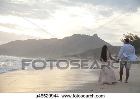 Picture - "Mid adult couple walking along beach, hand in hand, rear view". Fotosearch