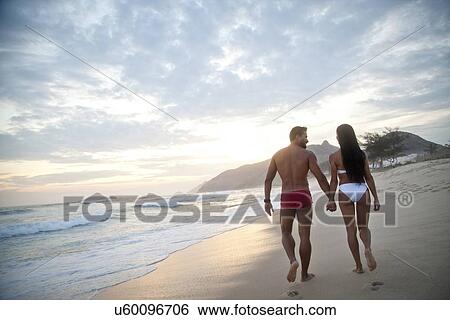 Stock Photograph - "Mid adult couple walking along beach, hand in hand, wearing swimwear, rear view". Fotosearch