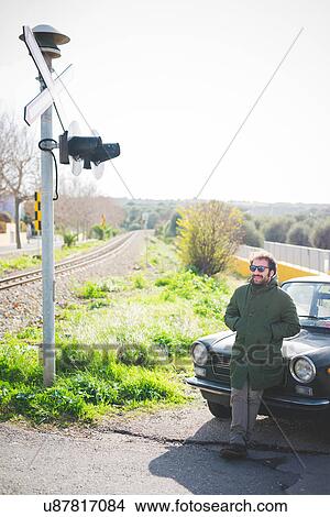 Picture - Mid adult man leaning against vintage car at railway crossing. Fotosearch