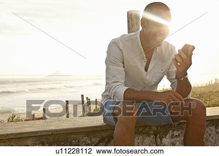 Stock Image - "Mid adult man reading smartphone texts at beach, Rio De Janeiro, Brazil". Fotosearch