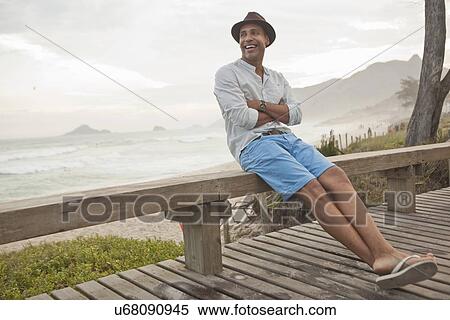 Stock Photography - "Mid adult man sitting on boardwalk fence, Rio De Janeiro, Brazil". Fotosearch
