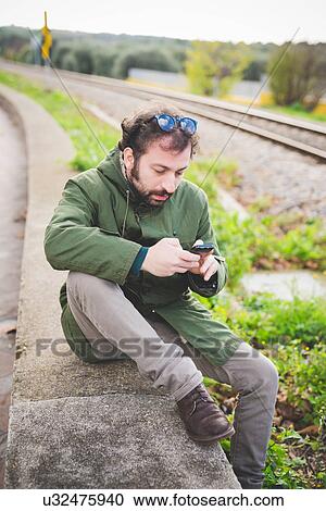 Mid adult man sitting on wall next to railway track using smartphone View Large Photo Image Stock Image - Mid adult man sitting on wall next to railway track using smartphone. Fotosearch