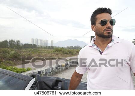 Stock Image - Mid adult man standing by convertible car by side of road. Fotosearch