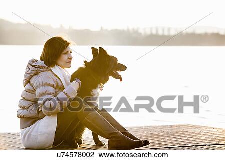 Mid adult woman and her dog sitting on sunlit lake pier View Large Photo Image Stock Image - Mid adult woman and her dog sitting on sunlit lake pier. Fotosearch