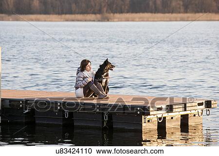 Stock Image - Mid adult woman and her dog sitting on lake pier. Fotosearch