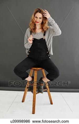 Mid adult woman sitting on stool wearing dungarees View Large Photo Image Stock Photograph - Mid adult woman sitting on stool wearing dungarees. Fotosearch