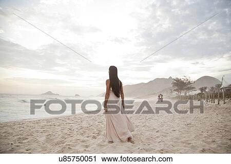Stock Image - "Mid adult woman standing on beach, rear view". Fotosearch