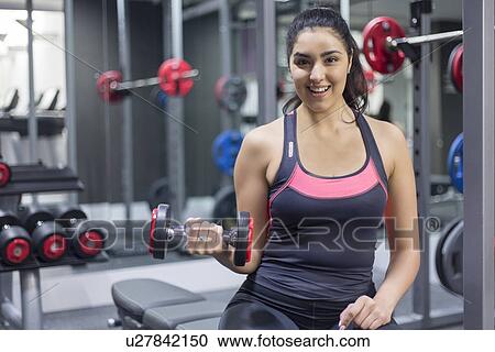 Portrait of a smiling young woman at gym holding dumbbell View Large Photo Image Stock Image - Portrait of a smiling young woman at gym holding dumbbell. Fotosearch