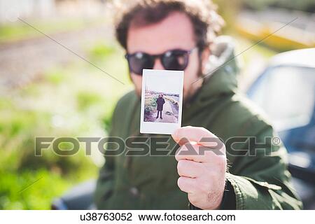 Stock Image - Portrait of mid adult man holding up an instant photograph of himself. Fotosearch