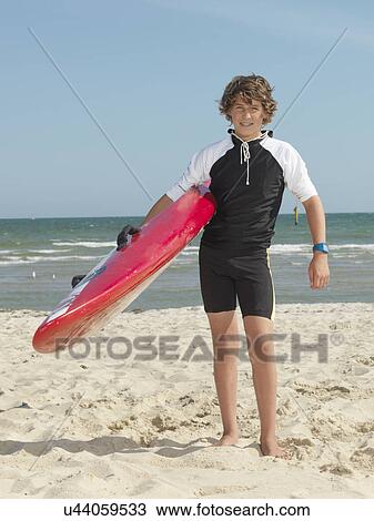 Stock Image - "Portrait of teenage boy nipper (child surf life savers) carrying surfboard, Altona, Melbourne, Australia". Fotosearch