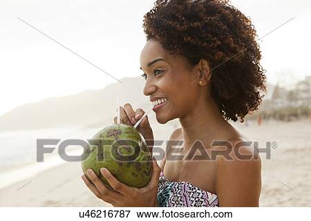 Stock Photo - "Portrait of young woman drinking coconut milk on beach, Rio De Janeiro, Brazil". Fotosearch