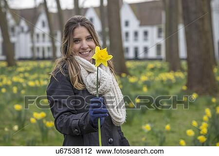 Stock Image - Portrait of young woman in spring park holding up daffodil. Fotosearch