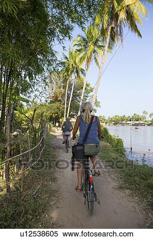 Stock Photography - "Rear view of two female tourists cycling on Mekong riverbank, Don Det, Laos". Fotosearch