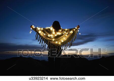 Picture - Rear view of woman wrapped in shawl with star fairy lights on beach at night. Fotosearch