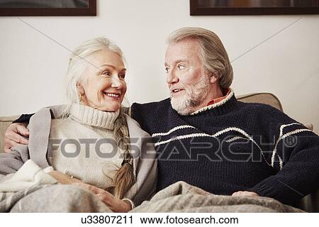 Stock Image - "Senior couple sitting on sofa, smiling". Fotosearch