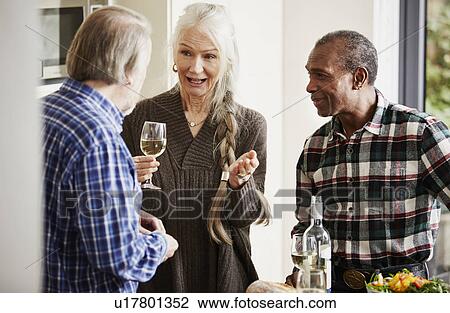 Stock Image - Senior friends talking in kitchen with wine. Fotosearch