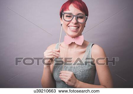 Stock Image - Studio portrait of confused young woman holding up spectacles in front of face. Fotosearch