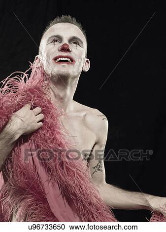 Stock Image - Studio portrait of young man in clown face paint with pink feather boa. Fotosearch