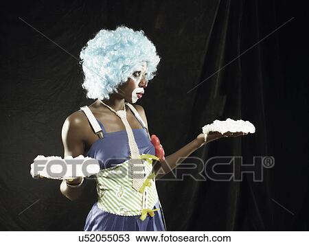 Stock Image - Studio portrait of young woman wearing clown face paint and blue wig holding meringues. Fotosearch