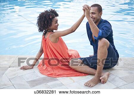 Stock Image - "Stylish couple sitting holding hands at hotel poolside, Rio De Janeiro, Brazil". Fotosearch