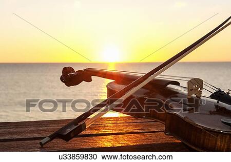Stock Image - Sunset over sea with violin and bow on bench. Fotosearch